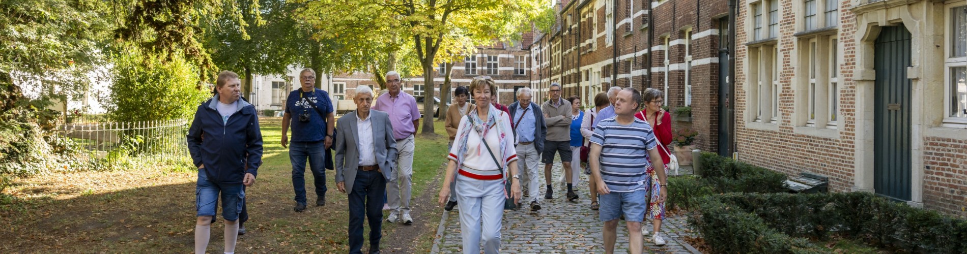 Groep mensen wandelt op een zonnige dag langs oude bakstenen huizen en bomen in een parkachtige omgeving.