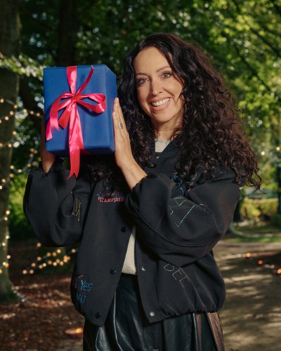 Vrouw met krullend haar lacht en houdt een blauw cadeau met rode strik vast in een bosrijke omgeving.