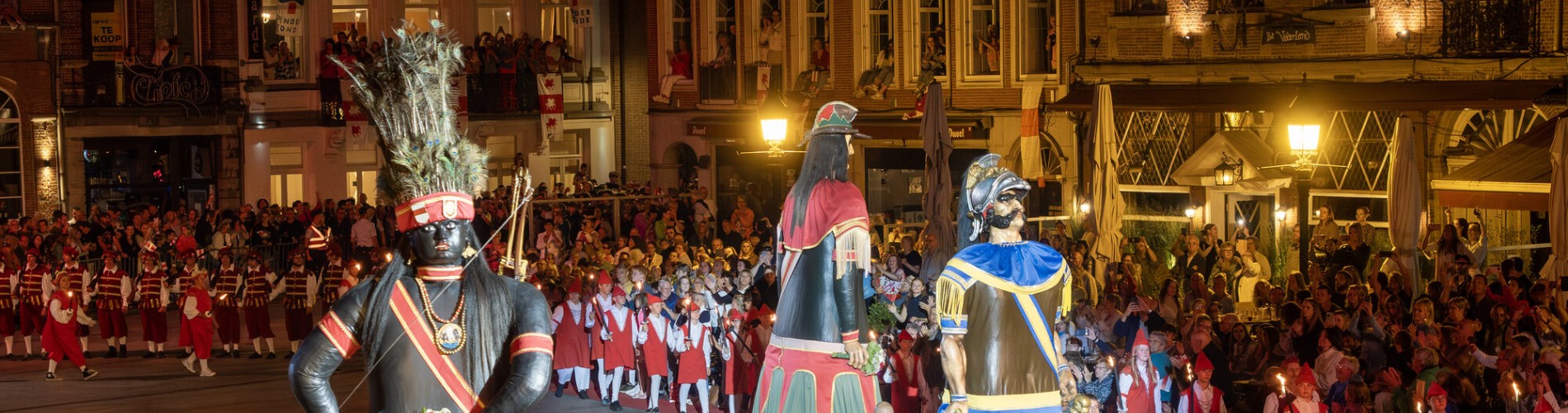Drie grote reuzenpoppen in kleurrijke kleding tijdens een nachtelijke parade met veel toeschouwers.