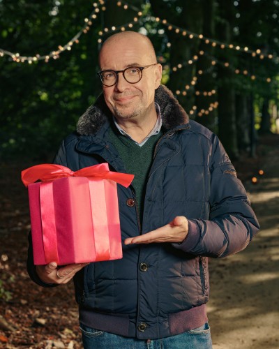 Man met bril en winterjas houdt een rood cadeau met strik vast in een bos met sfeerverlichting.
