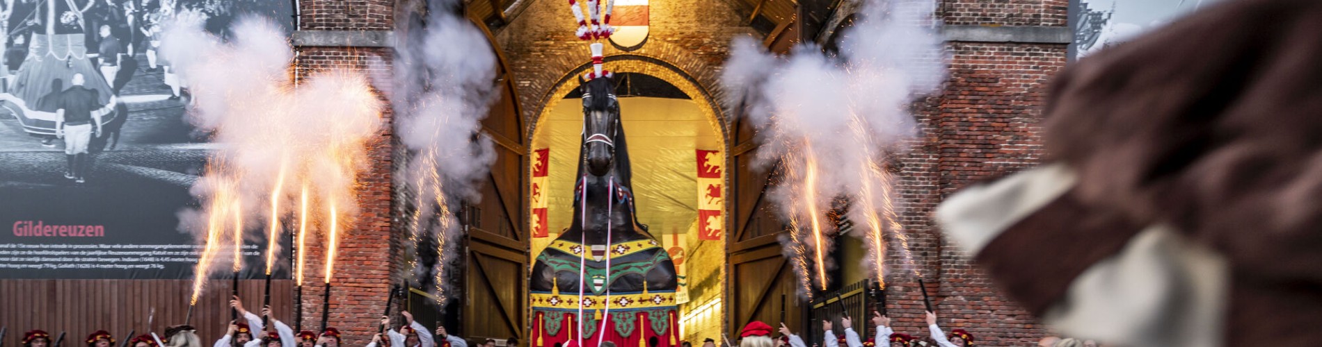 Traditionele optocht met reuzenfiguur en vuurwerk bij een grote poort, omringd door mensen in historische kleding.
