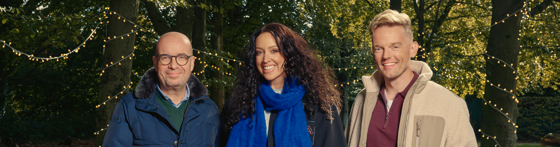 Drie mensen in warme kleding staan buiten op een pad met bomen en lichtjes in de avond.