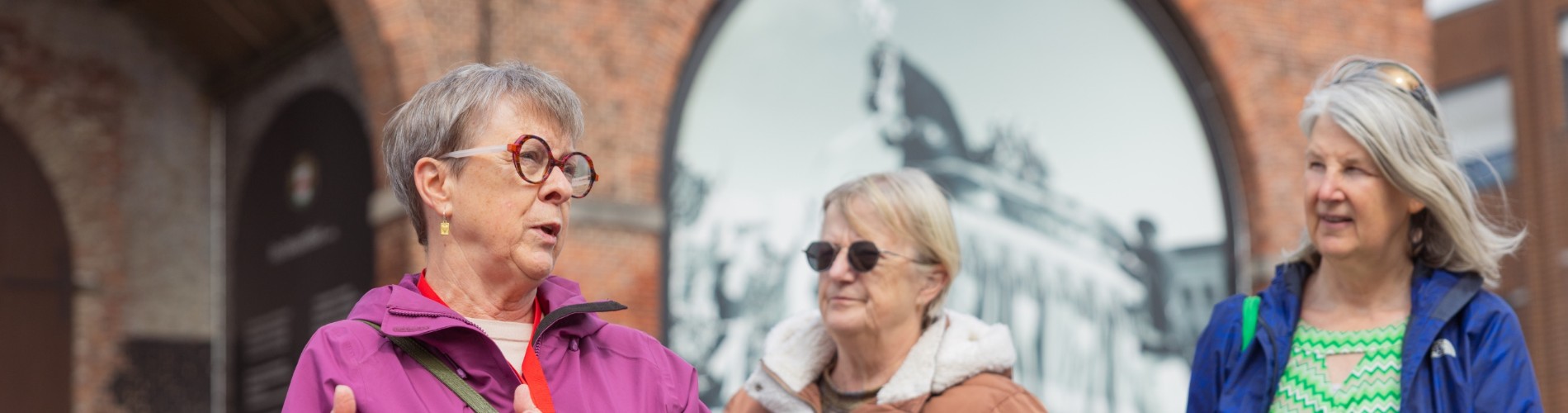 Drie oudere vrouwen in jassen staan buiten, één vertelt iets terwijl de anderen luisteren bij een historische muur.