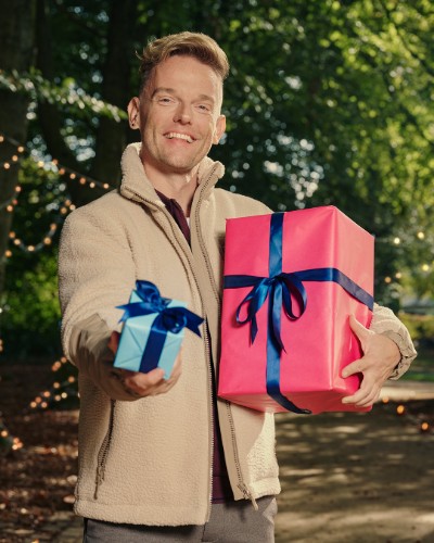 Man met beige jas houdt twee cadeaus vast, een grote roze en een kleine blauwe met blauwe linten, buiten in het park.